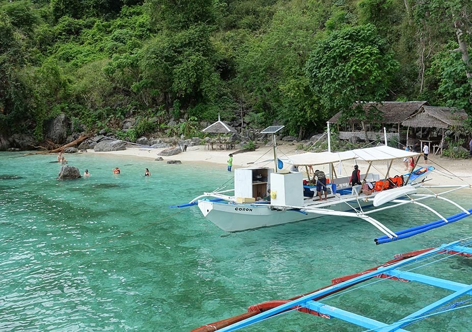 Traditional boats are parked near to a small beach on a limestone island covered in jungle, the water is clear blue.