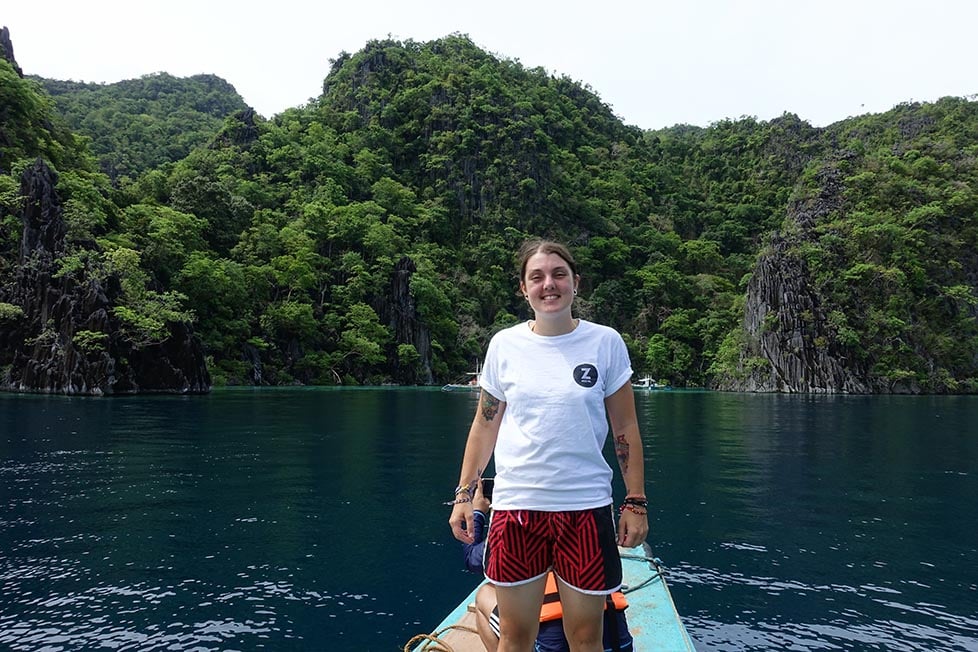 A person standing on the front of a boat with jungle covered cliffs in the background