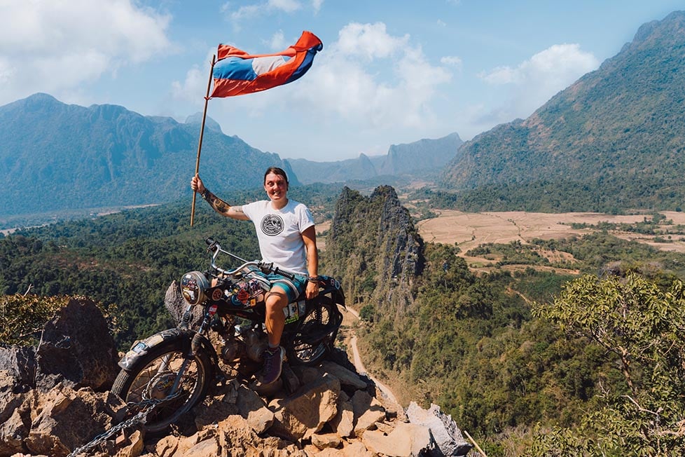 A person sitting on top of a motorbike at the top of a mountain in Vang Vieng. They have a Laotian flag in their hands and behind them are more jagged jungle covered limestone peaks.