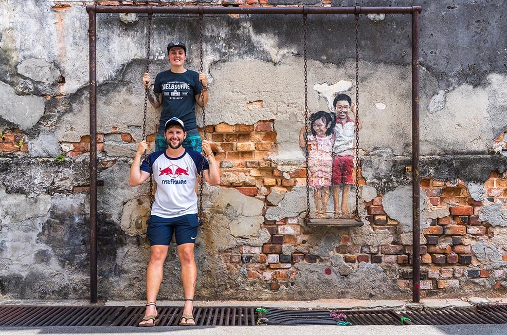 Two people standing on a swing next to some interactive street art in Penang, Malaysia.