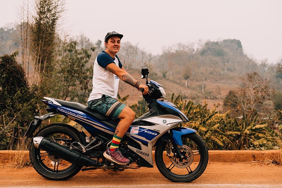 A person on a motorbike in Thailand with dry hillside and fields behind them.