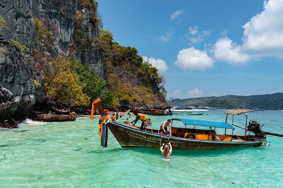 A traditional Thai longboat floats in turquoise waters off the side of a limestone cliff near Phuket.