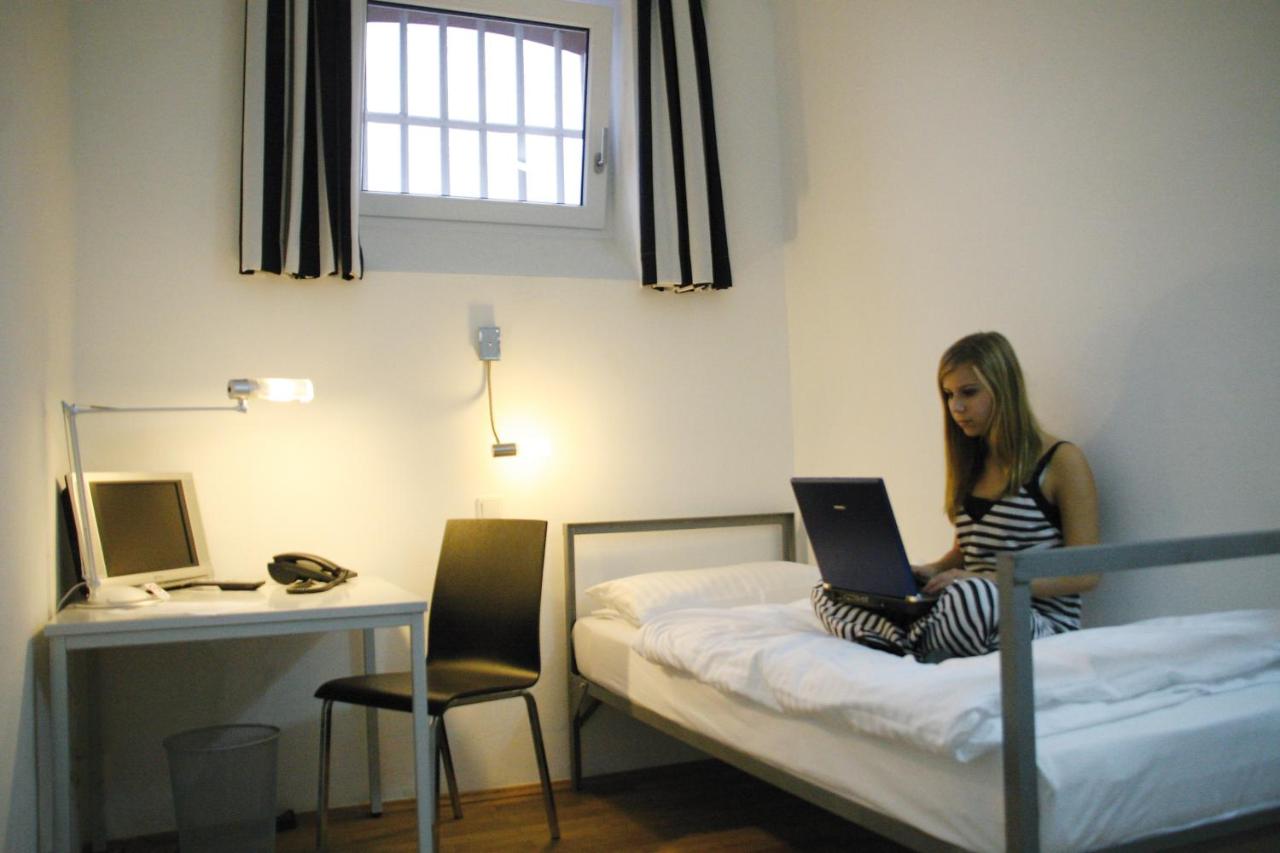A woman seated with her laptop on her lap in a prison hotel room.
