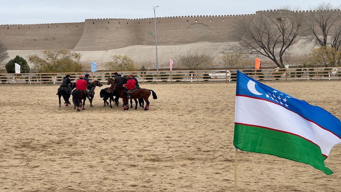 men sitting on horses getting ready to play kupkari in a sandy field beneath a fortress wall in uzbekistan with the uzbek flag waving