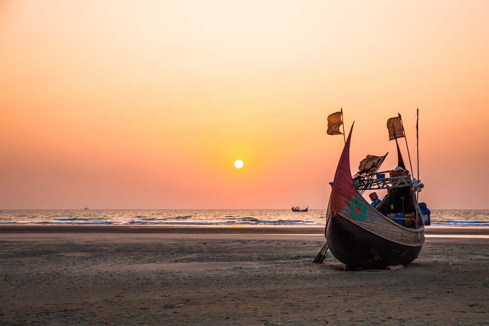 a wooden boat sitting on the beach during a pink and orange sunset in bangladesh