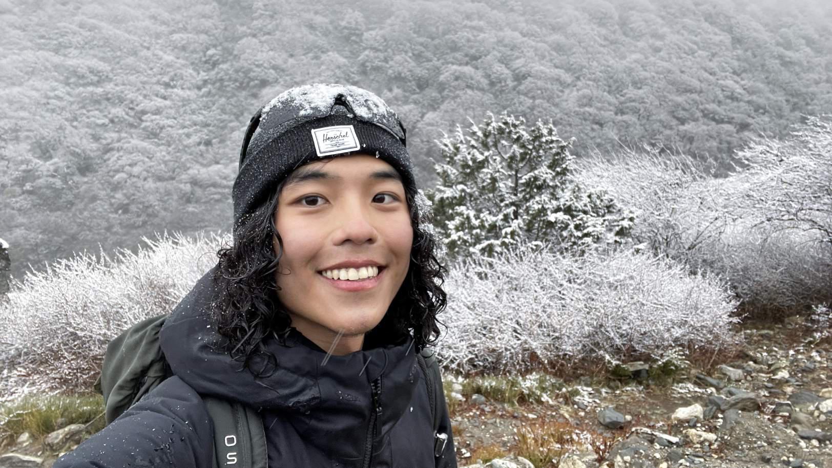 a guy hiking in nepal standing in front of snow covered bushes with snow coming down