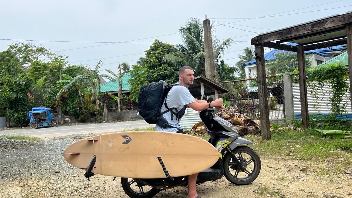 Joe on a scooter with a surfboard and Osprey Farpoint 40 - Hand lugagge only