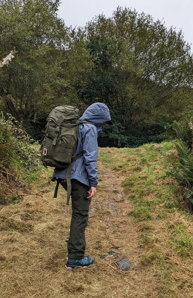 person facing away wearing a raincoat and camo coloured backpack walking up a grassy hill