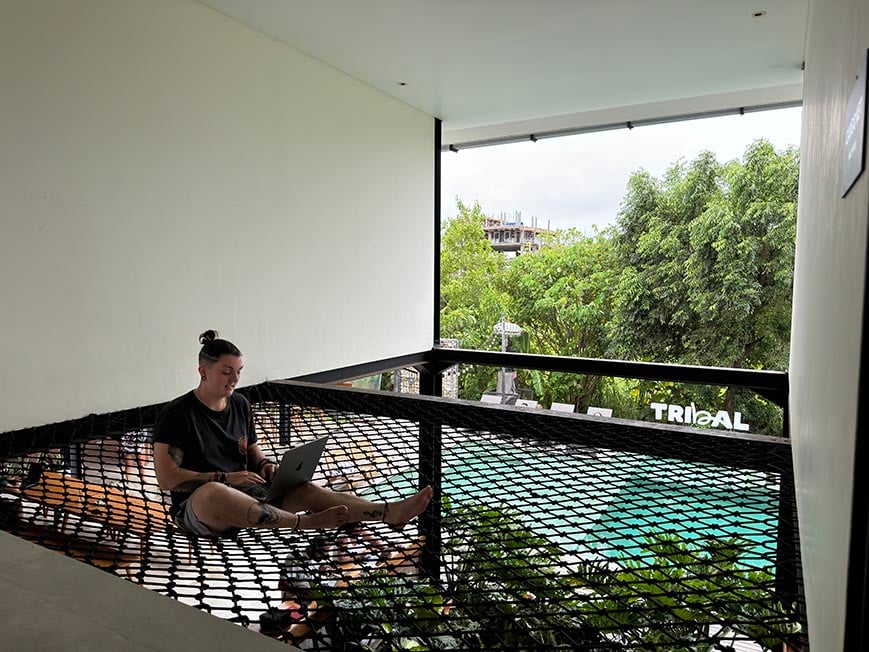 Nic working on their laptop on a cargo net at a hostel with a swimming pool in the background.