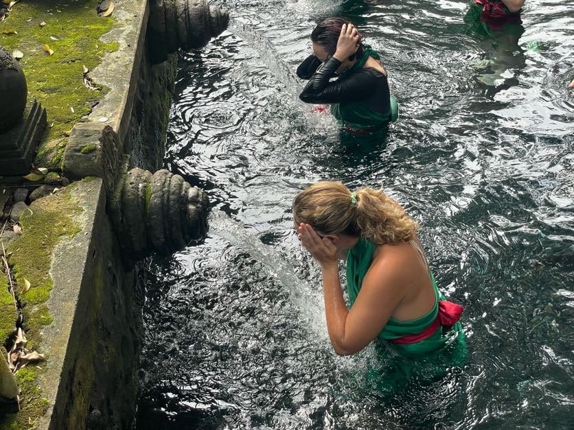 woman wearing a green robe in a pool washing her face with water pouring from a spout 