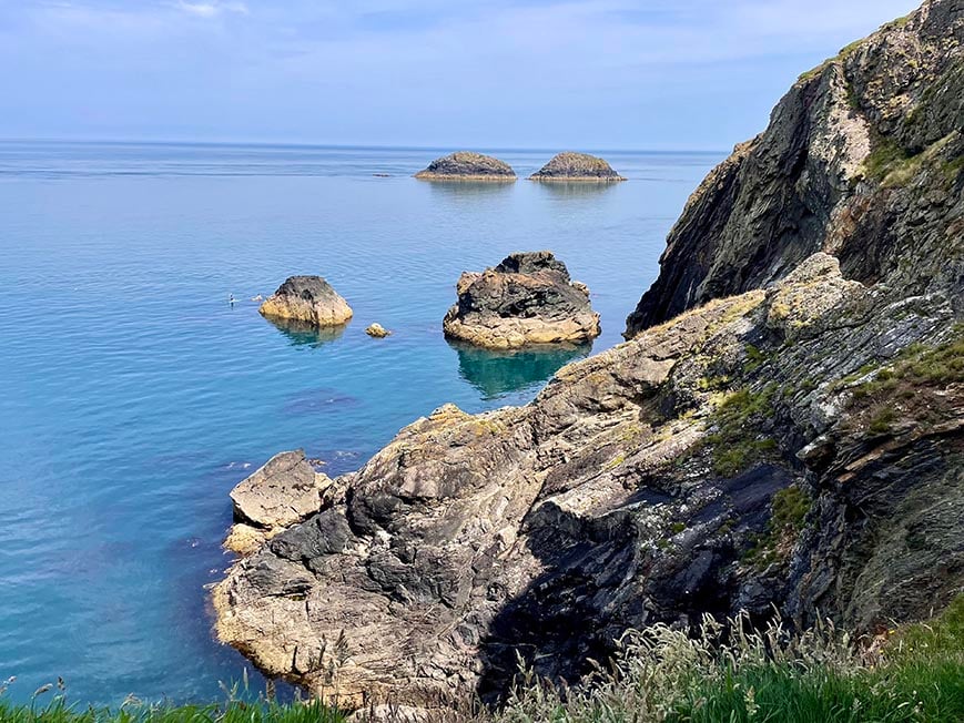 Sheer rugged cliffs and coastline with small rocky islands and clear blue water. 