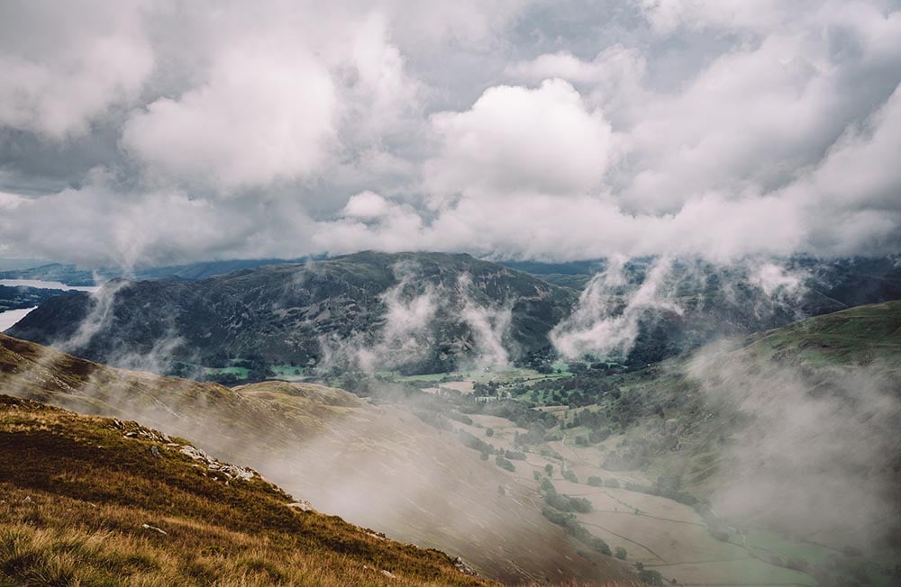 Mountains covered in trees and grass with wispy clouds blowing through the valleys.