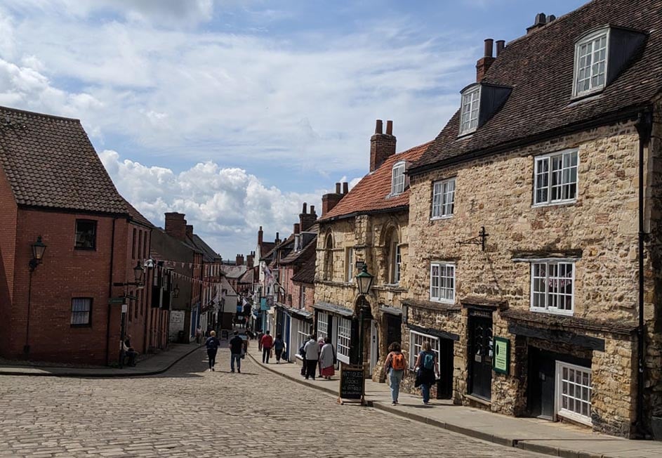A wide street in Lincoln, UK, bordered by old stone houses and people walking along the sidewalk