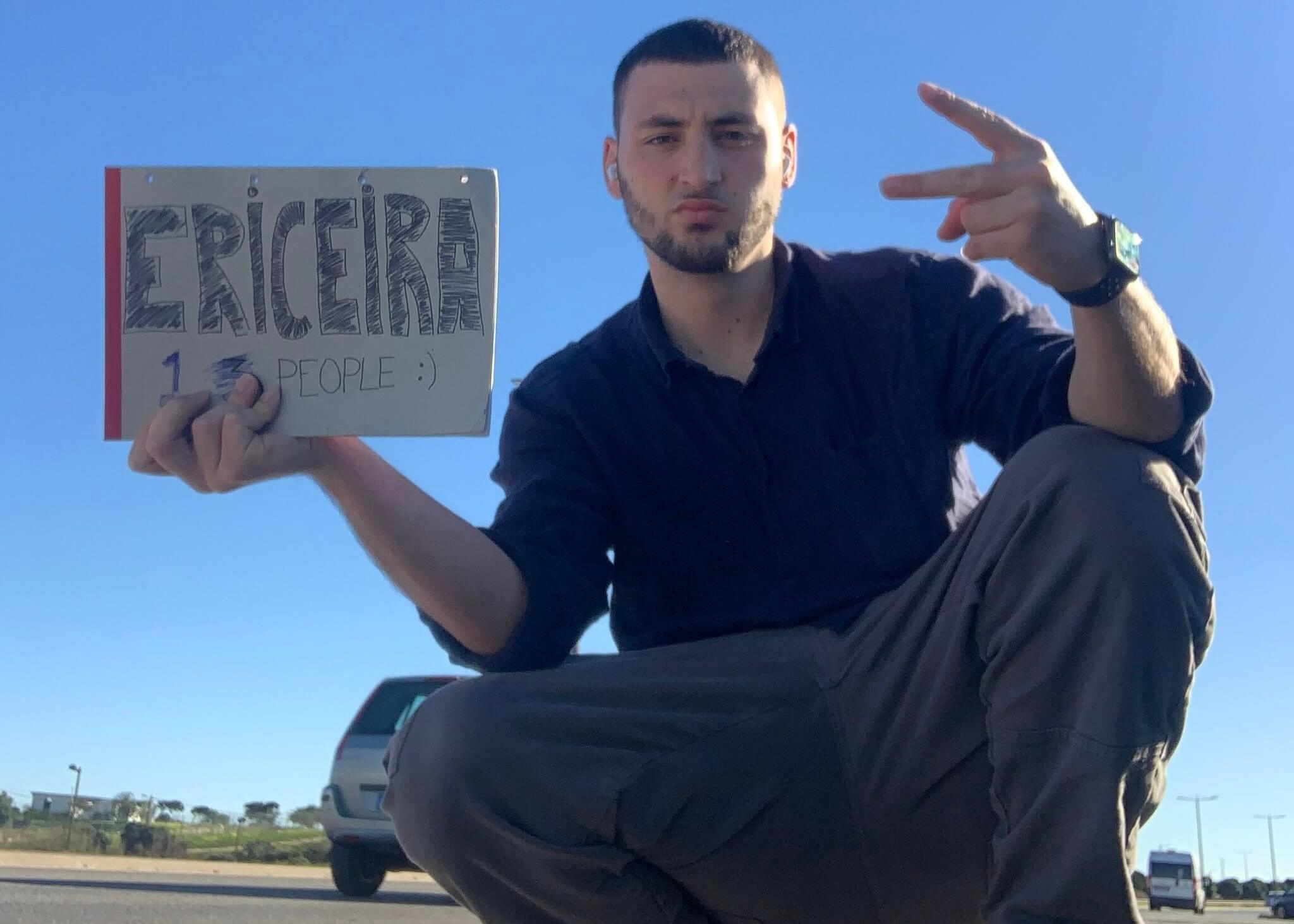 Man crouching by the side of a road with a cardboard hitchhike sign for Ericeira