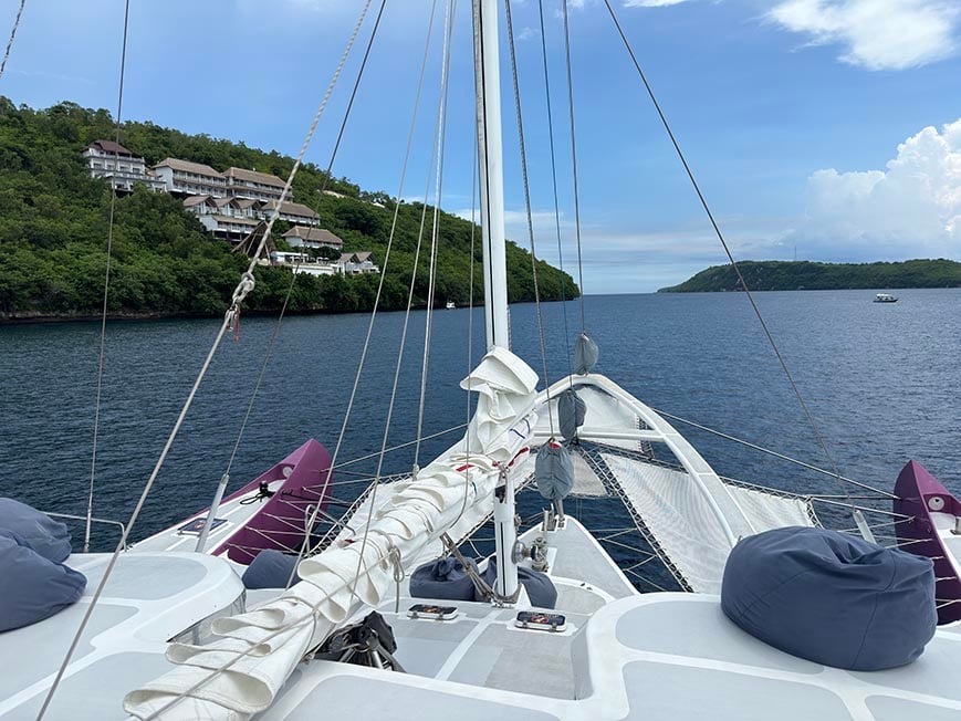 The front of a large boat in waters off the coast of jungle covered islands in blue waters with blue sky with a few clouds in the background.