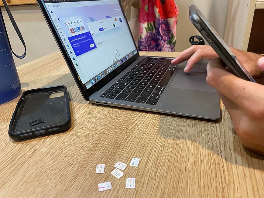 A pile of plastic sim cards from different providers on a table next to an open laptop and phone case and a persons hand with a phone in it.