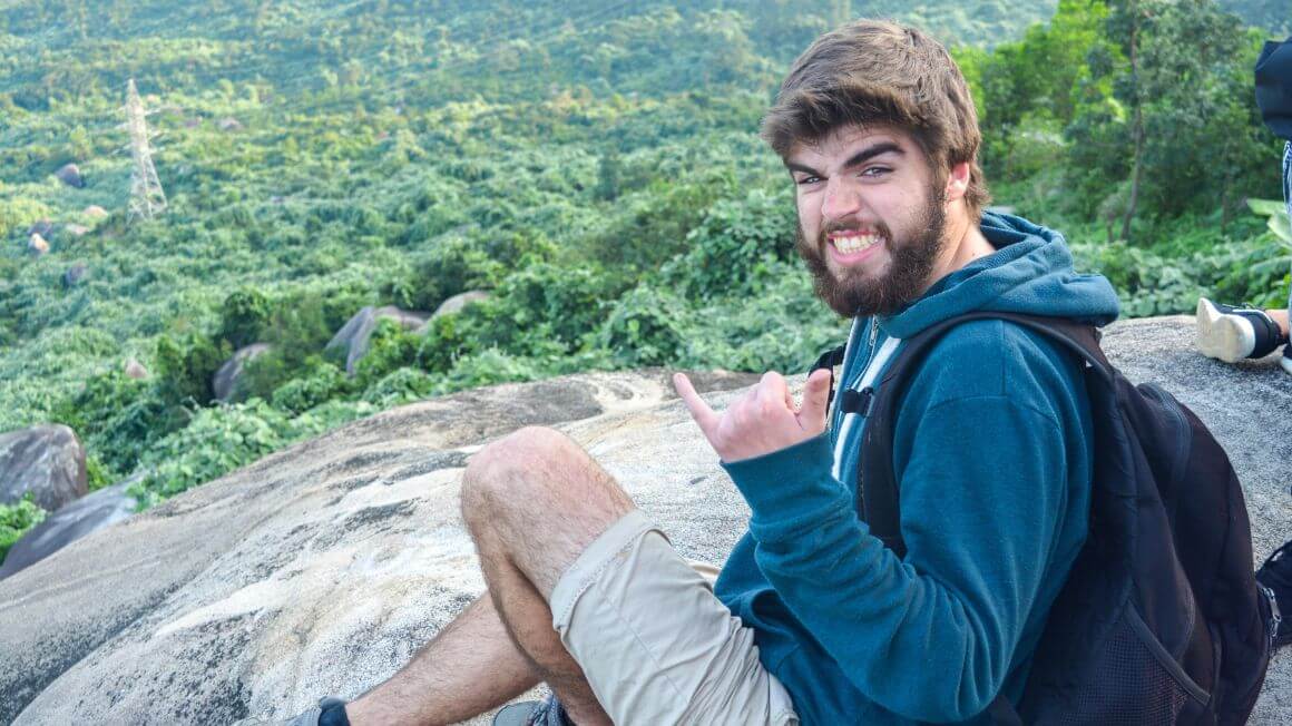 Tomas sat on a rock on a hill smiling awkwardly making a cowabunga hand symbol