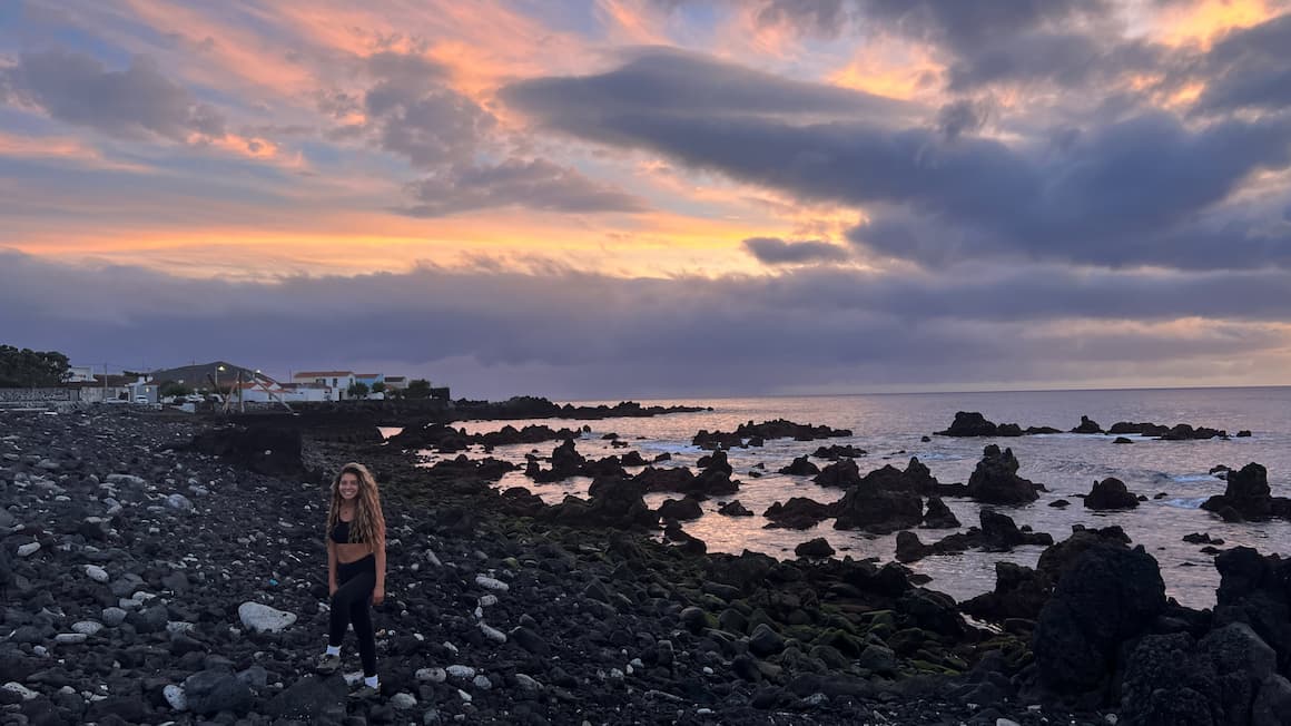 amanda smiling in front of a volcanic black rock beach as the sun set and the sky is orange and purple, on pico island in the azores, portugal 