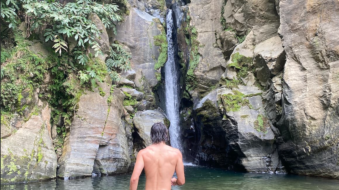 a guy facing a waterfall with large rocks surrounding it in sao miguel island, the azores, portugal