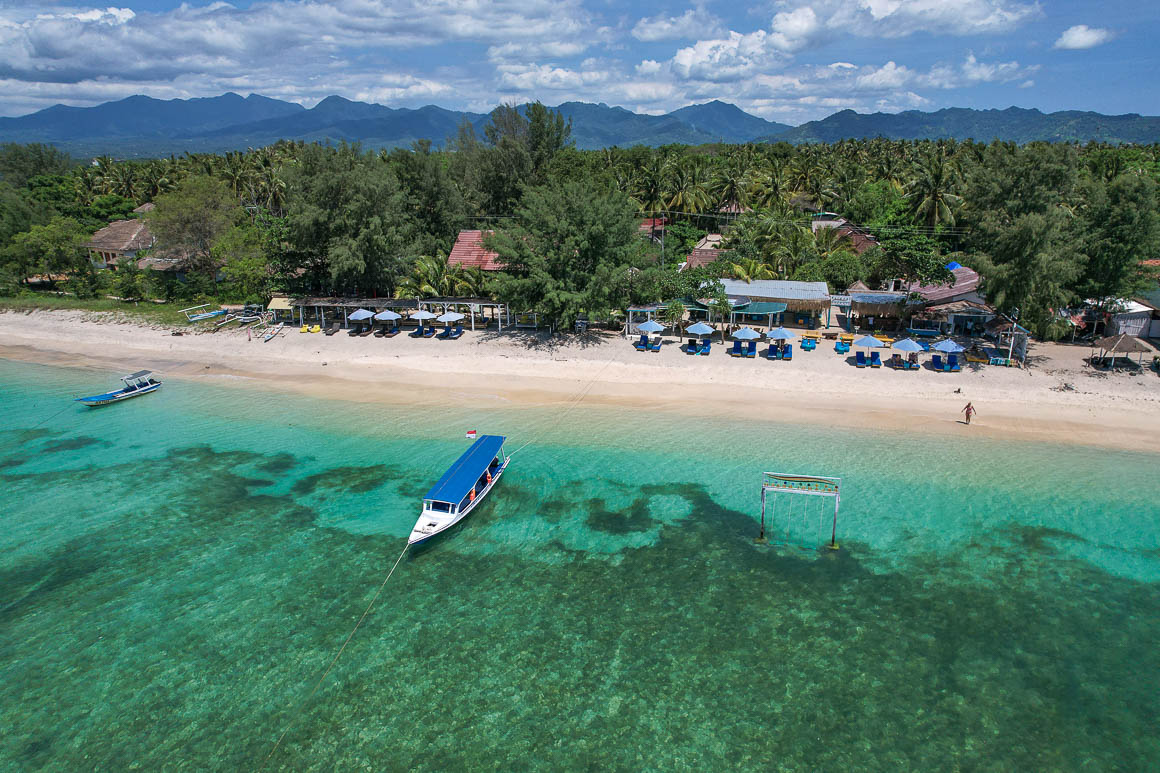 drone shot of boats and a swing at a turquoise colored beach in gili air with the mountainous landscape of north lombok island rising up in the distance