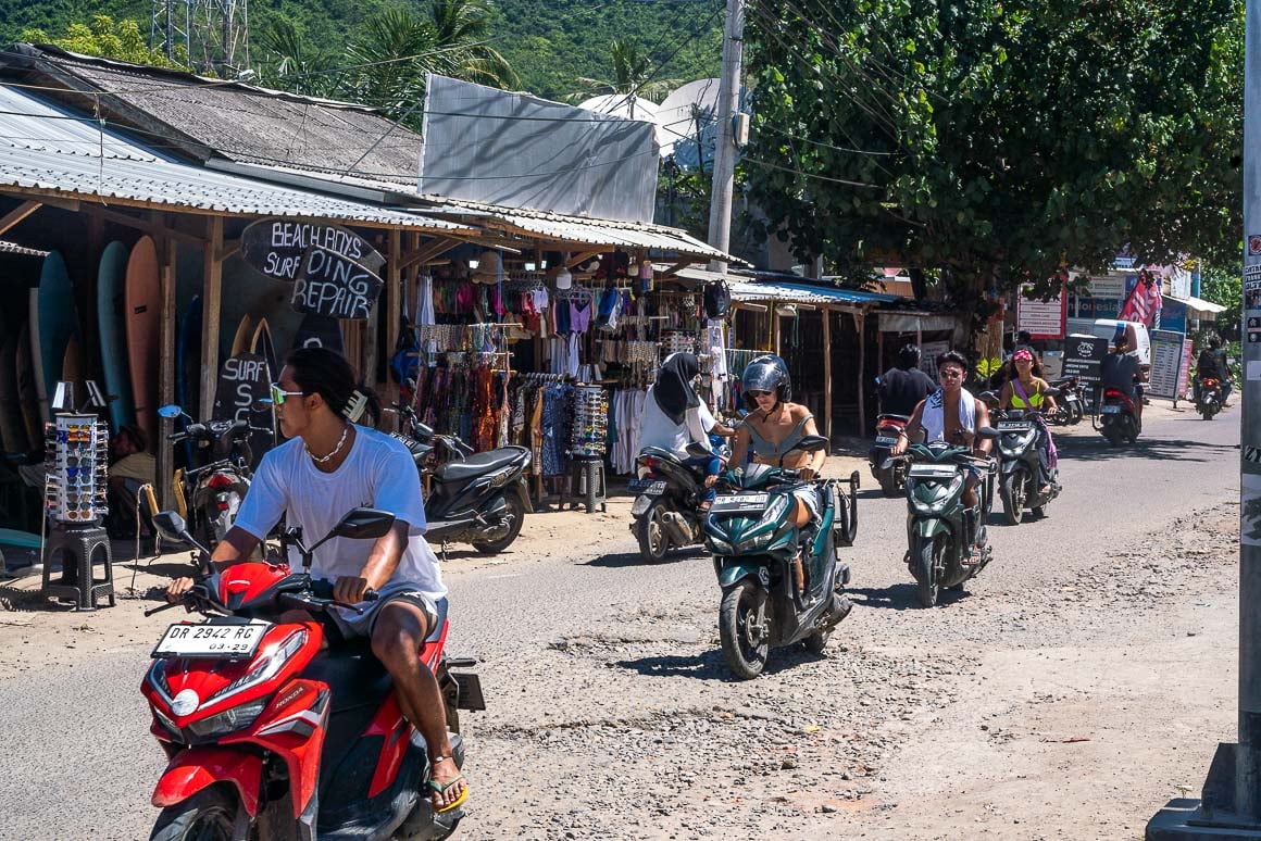 backpackers and surfers riding on scooters past clothing and surfboard rental shops in Kuta Lombok on a sunny day