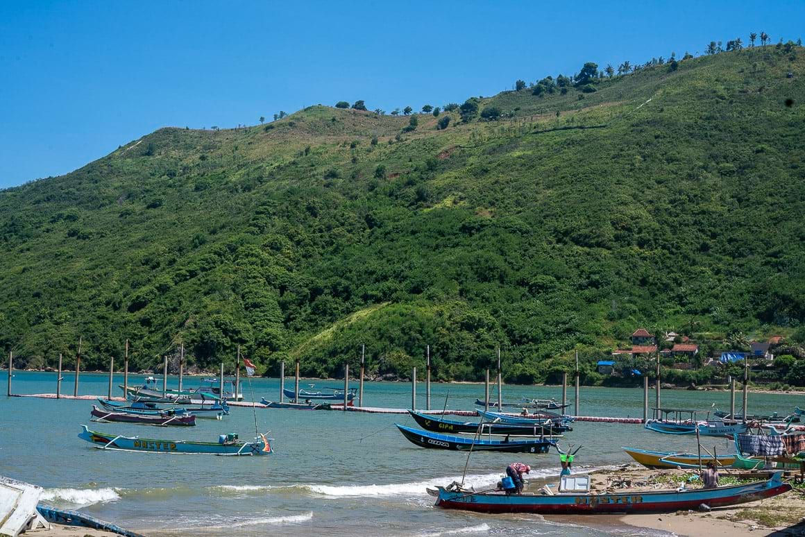 blue boats floating on almost clear turquoise water with a lush forested ridge towering behind it in kuta Lombok