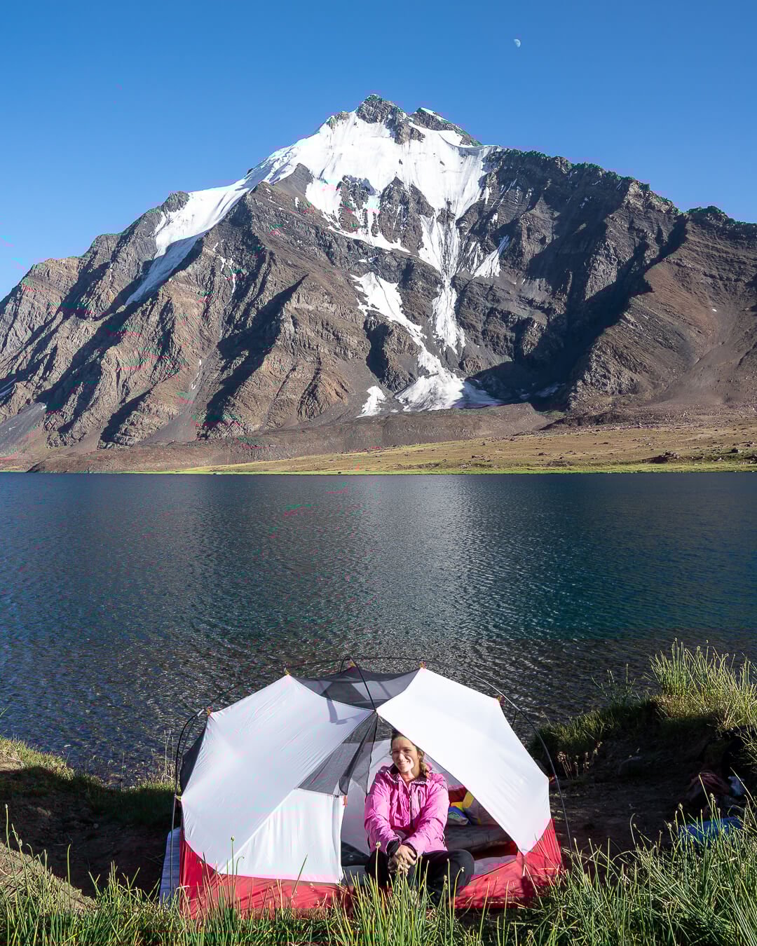 a girl sitting inside of a white and red tent in front of a huge blue alpine lake and snow capped mountain peak