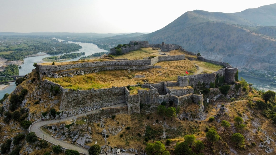 stone castle with green fields and a red flag overlooking a river in shkoder albania
