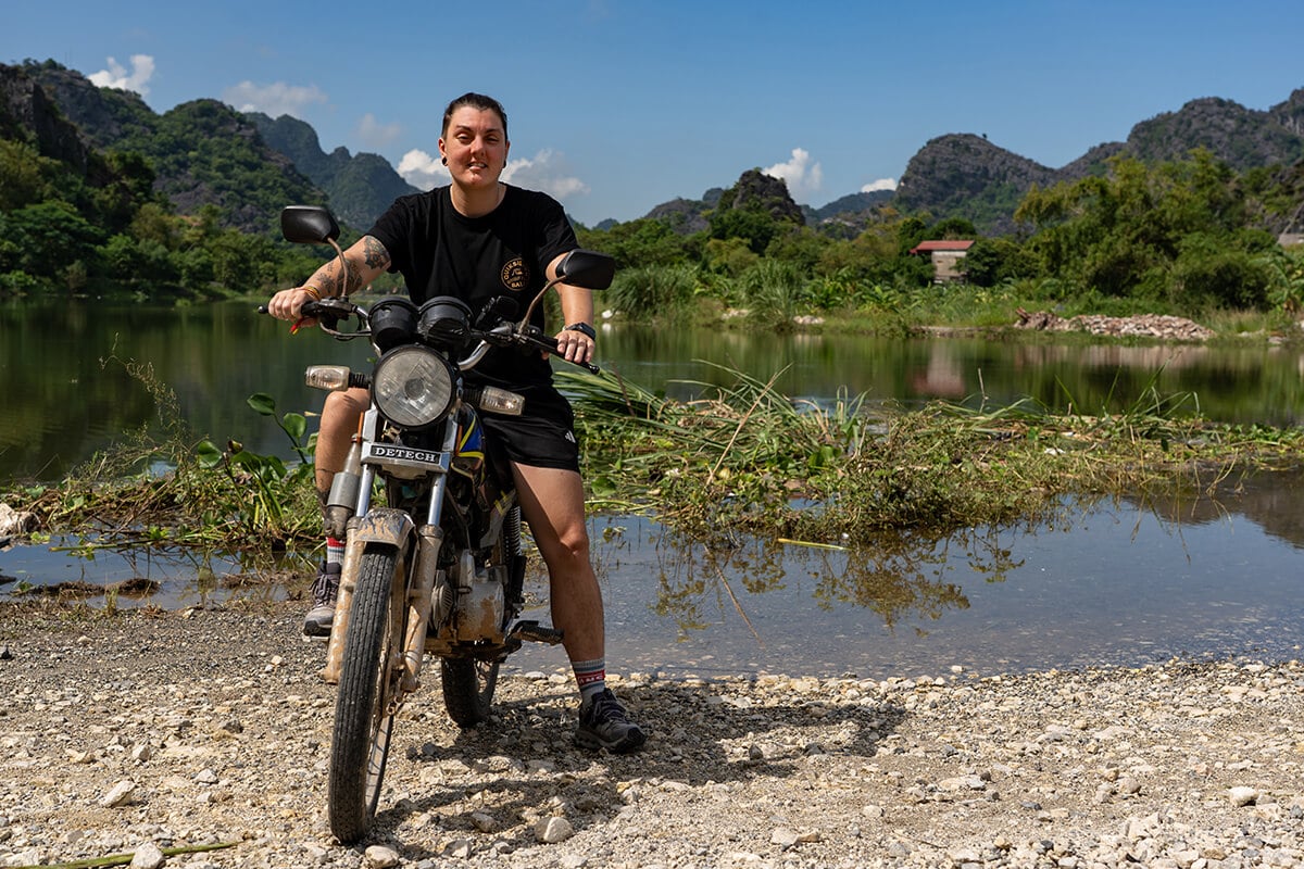 Nic being an absolute badass on their motorbike in Vietnam near Ninh Binh with mountains and a lake behind them.