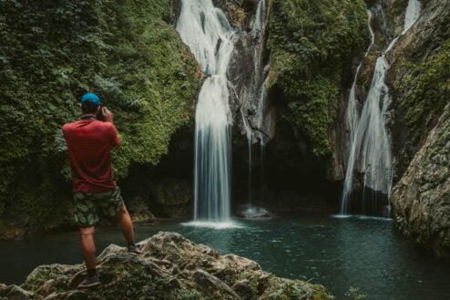 A person standing on a rock with a camera taking a photo of a waterfall