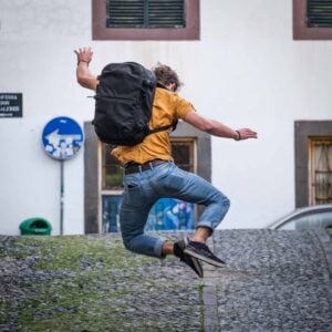 A person wearing a backpack is jumping into the air and clicking his feet along a cobbled road