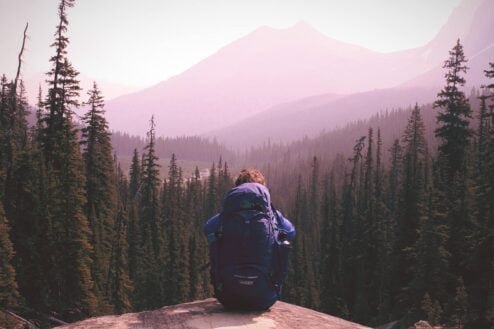 a person sits with her backpack facing a beautiful mountain range