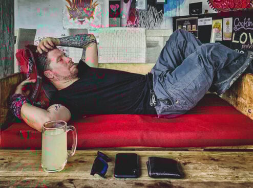 Will lies down on a make shift couch with a bag as a pillow with his phone, wallet, sunglasses and a drink next to him