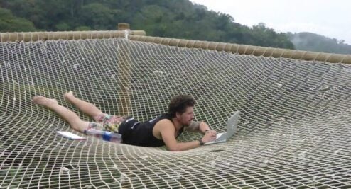 Will is laid down on his front on a net whilst working on his laptop