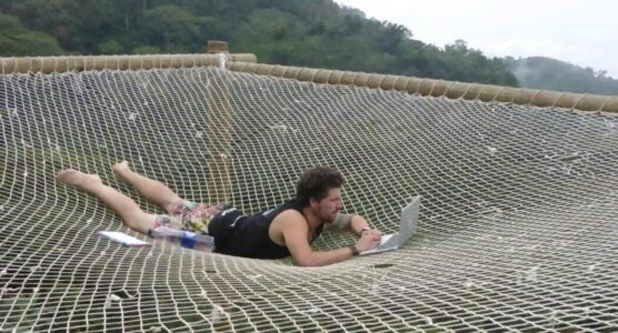 Will is laid down on his front on a net whilst working on his laptop