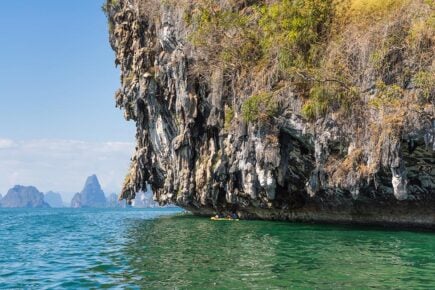 Two people kayak under an overhanging limestone cliff in Thailand with more peaks jutting from the sea in the distance.