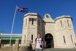 A person standing in front of Fremantle Prison in Western Australia. Behind them are the large stone entrance gates and an Australian flag.