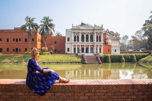 a girl in a blue dress sitting on a brick wall overlooking a canal and a white building in bangladesh