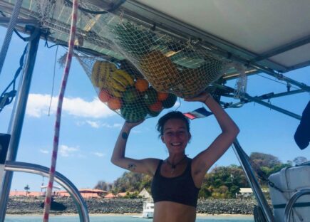 A girl is smiling on a sailboat while holding up bags of fruit.